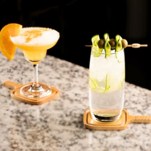 Two cocktails on a marble bar: one with an orange slice, the other with olives and cucumber garnish.