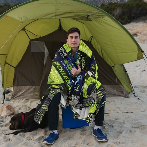 Man sitting on a cooler wrapped in a blanket at a beach campsite with a tent.