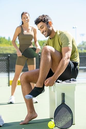 Two people on a pickleball court, one sitting and adjusting footwear, the other standing with a paddle.