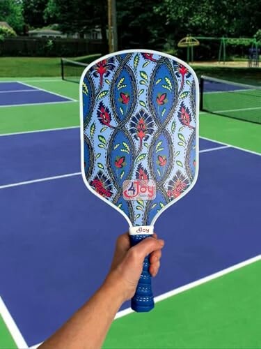 Hand holding a colorful pickleball paddle on a tennis court.