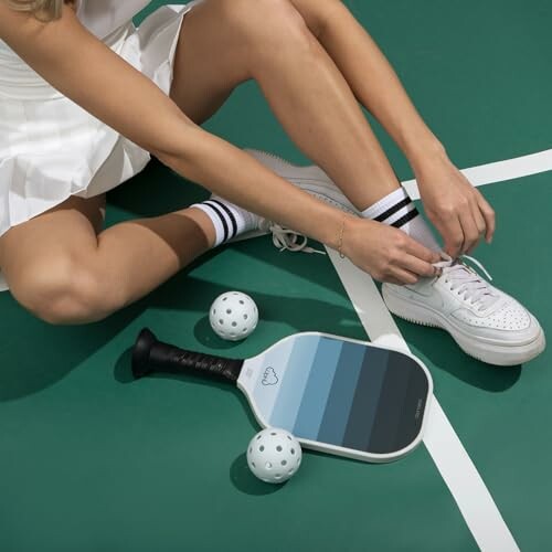 Person tying shoes next to a pickleball paddle and balls on a court.