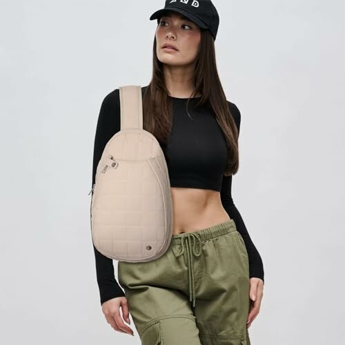 Woman in black top and green pants wearing a crossbody bag and cap.