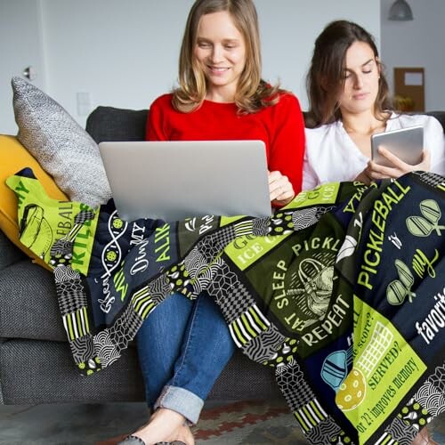 Two women sitting on a couch with a pickleball-themed blanket, using a laptop and a tablet.